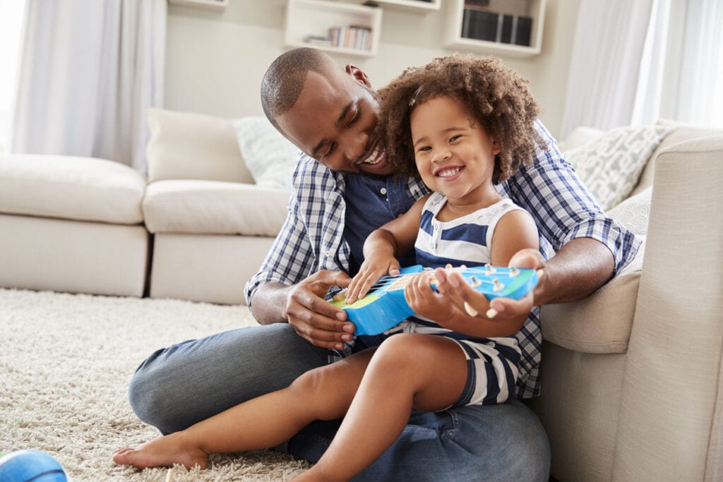 Toddler daughter sits on dad's knee playing ukulele at home