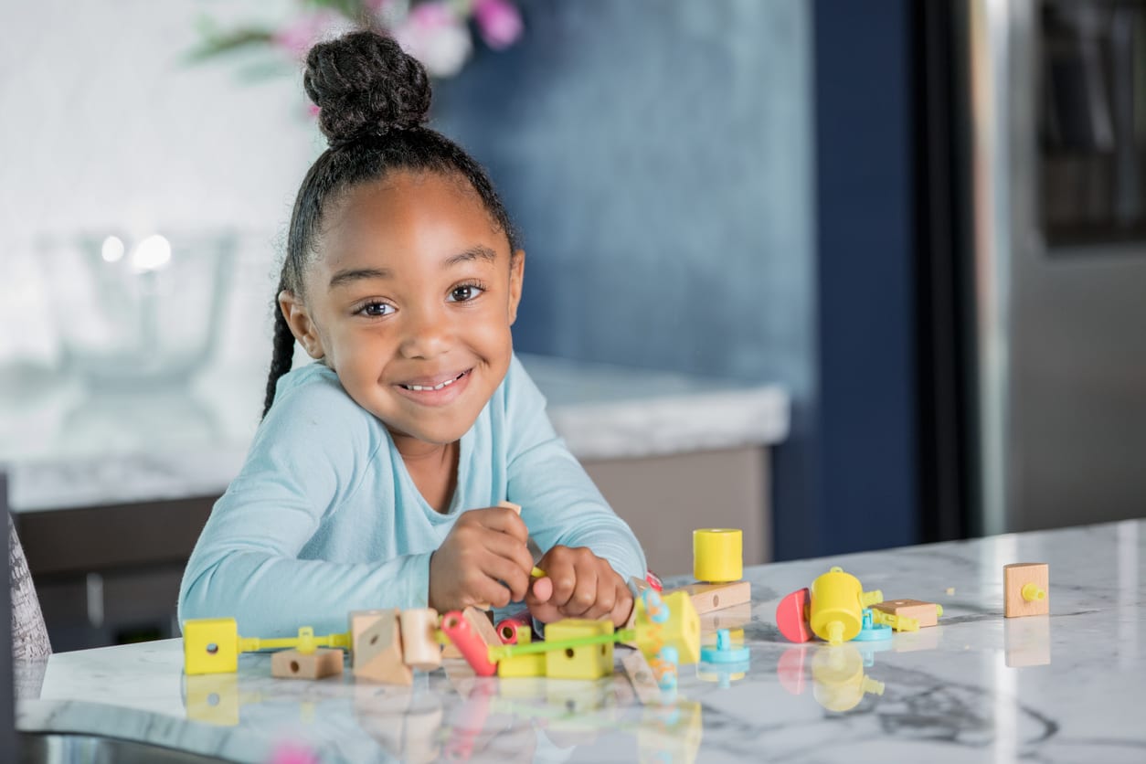 Adorable African American elementary age little girl is using a STEM educational toy at home in modern kitchen. She is looking at the camera and smiling.