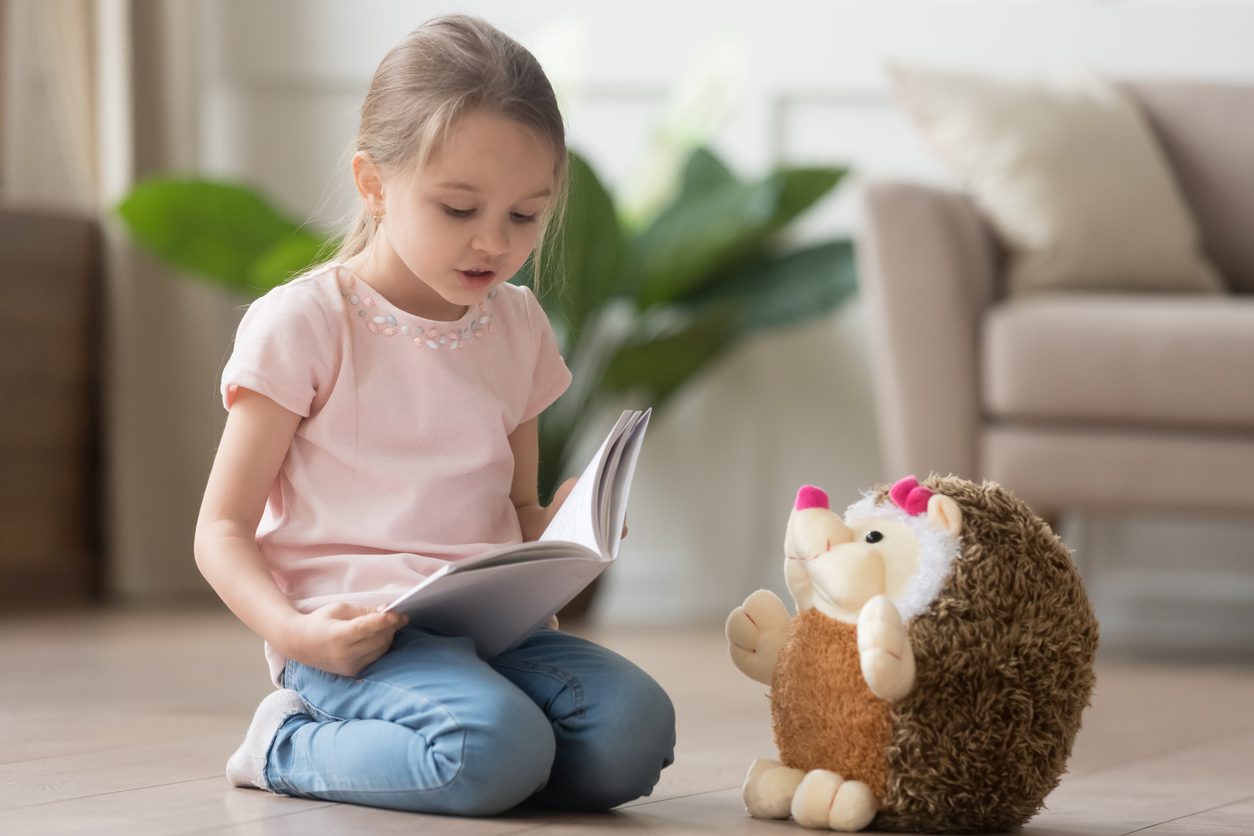 Cute smart little kid girl playing alone reading story to fluffy hedgehog sitting on warm floor at home, funny creative preschool small child holding book teaching toy, children imagination education