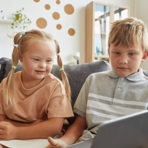 Portrait of cute girl with down syndrome smiling while looking at laptop screen and sitting on sofa with brother at home