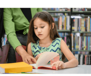 Little brunette girl is being instructed by a woman in a green shirt as they look at a book together.