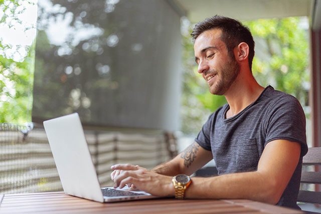 Young man in a gray tshirt works outside on his laptop. He is a professional teletherapist working for Connect Teletherapy.