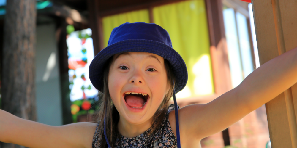 Smiling little girl with Down syndrome Trisomy 21 wears a blue bucket hat and spreads her arms wide