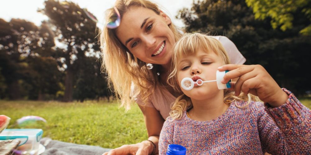 Cute little girl blowing soap bubbles while sitting with her mother outdoors. Mother and daughter having fun at picnic in park.