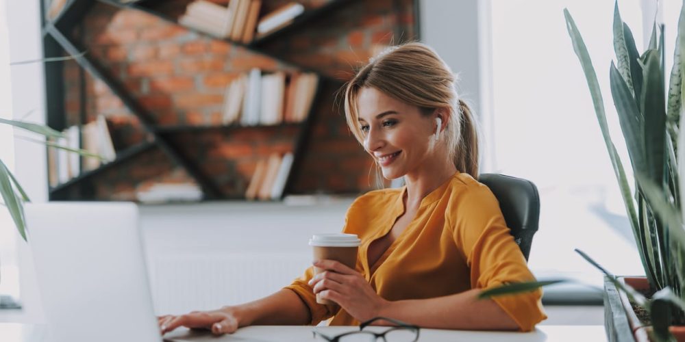 Blonde woman in an orange blouse having a carton cup of coffee in her hand and wearing wireless earphones while working on the laptop